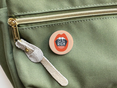 The Eat the Rich Pinback Button by Onderkast Studio, featuring bold red lips and a political message, is attached to the front pocket of a green bag with a gold zipper and beige suede pull for a striking statement.