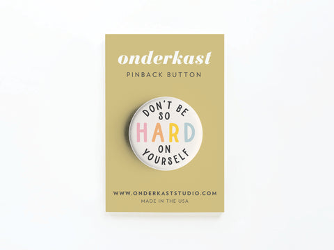The Don’t Be So Hard On Yourself Pinback Button by Onderkast Studio features “DONT BE SO HARD ON YOURSELF” with “HARD” in colorful letters, on a tan card with onderkast PINBACK BUTTON and www.onderkaststudio.com MADE IN THE USA.