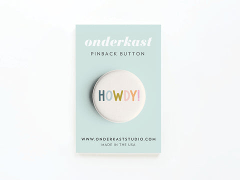 The Howdy! Pinback Button by Onderkast Studio features vibrant colors on a light blue card, with the brand name and website printed above and below. Proudly made in the USA.