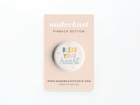 The Bless Your Heart Pinback Button by Onderkast Studio features colorful lettering and heart illustrations, adding Southern sass. Displayed on a pale pink card, it’s a playful accessory from the Onderkast Studio collection.