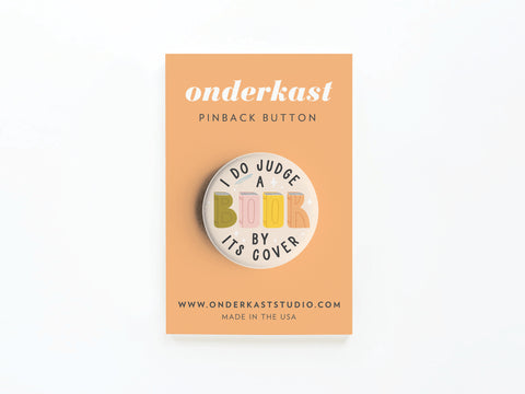 A round pinback button on a peach backing card reads I DO Judge a Book By Its Cover, with colorful lettering. Printed in the USA by Onderkast Studio. Text includes “Onderkast Pinback Button” and onderkaststudio.com.