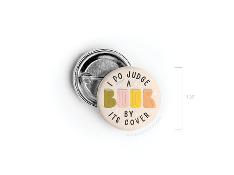 The Onderkast Studio “I DO Judge a Book By Its Cover” pinback button features colorful lettering on BOOK, measures 1.25 inches, and is printed in the USA. The round badge is shown on a white background with its size for reference.