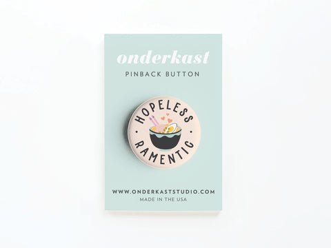 The Hopeless Ramentic Pinback Button by Onderkast Studio features a bowl of ramen and playful text, displayed on a mint green card. Website info and Made in the USA are shown below.