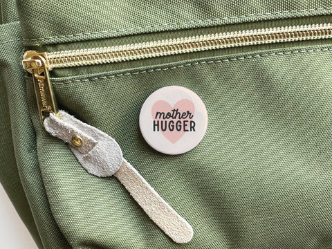 The Onderkast Studio Mother Hugger Pinback Button, displayed on a green fabric bag with a gold zipper and suede pull, features “mother HUGGER” in front of a pink heart—perfect as a charming Mother’s Day gift.