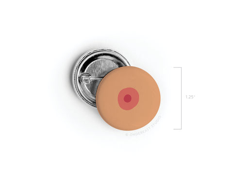 The Boob Pinback Button – Medium by Onderkast Studio is a 1.25-inch round pin featuring a light brown nipple and areola design, shown with its pin clasp back. Celebrate body positivity and self-love with this unique accessory!.