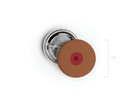 Celebrate self-love with the Onderkast Studio Boob Pinback Button – Dark, a 1.25-inch round button featuring a brown base and dark red center, resembling a nipple. Shown next to its pin-back side for reference.