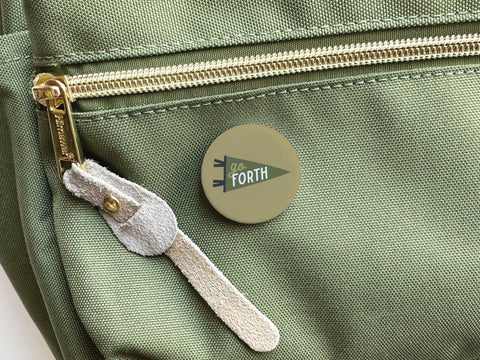 The Onderkast Studio Go Forth Pinback Button adds an adventurous flair to the front pocket of a light green backpack with a gold zipper and suede pull. This green button features Go Forth on a pennant design.