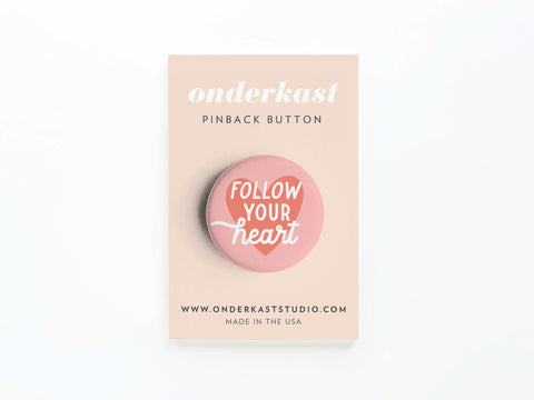 The Follow Your Heart Deep Pinback Button by Onderkast Studio is pink with white text, displayed on a beige onderkast PINBACK BUTTON card featuring whimsical details, the website, and Made in the USA at the bottom.
