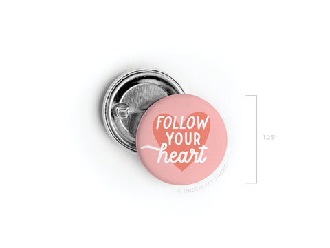 The Follow Your Heart Deep Pinback Button by Onderkast Studio is a 1.25-inch round pink button with FOLLOW YOUR heart in white text over a heart design, shown front and back.