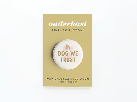 The In Dog We Trust Pinback Button by Onderkast Studio features bold text on a round button, mounted on a tan card displaying the brand and website. MADE IN THE USA appears at the bottom—perfect for dog lovers.