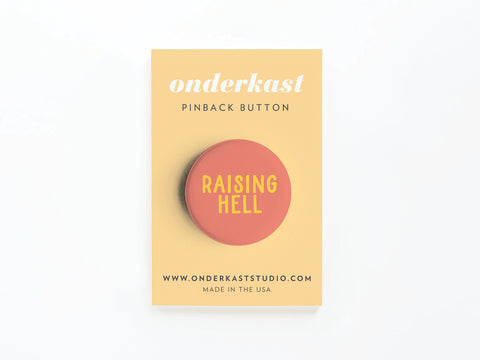 The Raising Hell Pinback Button by Onderkast Studio features bold yellow text on a round button, mounted on a tan card labeled onderkast PINBACK BUTTON with website and MADE IN THE USA details—a rebellious addition to any collection.
