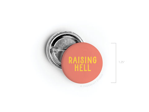 The Onderkast Studio Raising Hell Pinback Button is a 1.25-inch round accessory with a coral pink background and bold yellow text, featuring a metal pin clasp—perfect for adding attitude to any style.