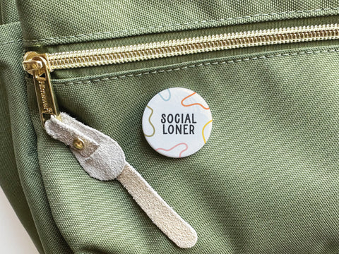 An Onderkast Studio Social Loner Pinback Button, featuring a light background with abstract colorful lines, is pinned to the front pocket of a zippered light green bag with a beige suede pull tab.