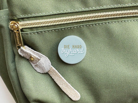 A green fabric bag with a gold zipper and suede pull tab is shown with the Die Hard Tryhard Pinback Button by Onderkast Studio—a light blue round pin reading DIE HARD tryhard in white and peach, made with high-quality craftsmanship.