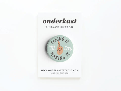 The Faking It ’Til I’m Making It Pinback Button by Onderkast Studio features a crossed fingers hand on a vibrant display card labeled “onderkast PINBACK BUTTON”—a fun way to show perseverance and determination.
