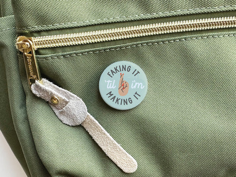 The Onderkast Studio Faking It ’Til I’m Making It Pinback Button, featuring bold colors and a crossed fingers design, rests on a green fabric bag—a fun reminder to keep persevering.