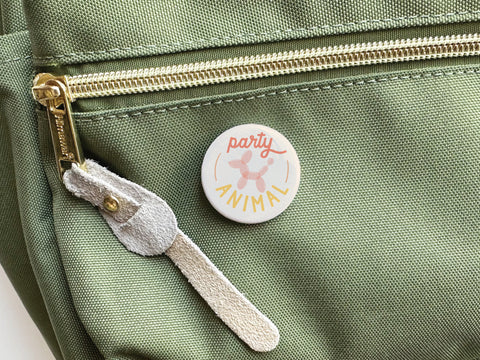 The Onderkast Studio Party Animal Pinback Button, featuring a colorful pink balloon dog design, is shown resting on a green fabric bag with a gold zipper and tan suede pull.