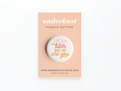 The Life’s a Bitch But So Are You Pinback Button by Onderkast Studio features pink and orange script on a peach card, is MADE IN THE USA, crafted with high-quality production, and includes website info at the bottom.