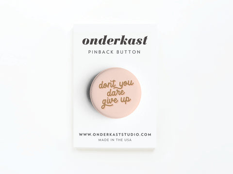 The Don’t You Dare Give Up Pinback Button by Onderkast Studio features elegant cursive font, comes attached to a branded card stating www.onderkaststudio.com and MADE IN THE USA, emphasizing its high-quality production.