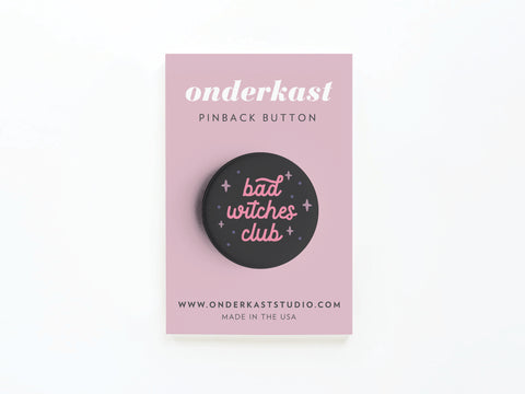 The Bad Witches Club Pinback Button by Onderkast Studio is a round black button with pink text and sparkles, displayed on a branded card. Made in the USA—perfect for your mystical sisterhood!.