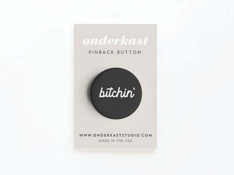 The Bitchin’ Pinback Button by Onderkast Studio features “bitchin’” in white on black, attached to a beige card labeled “onderkast PINBACK BUTTON,” with website info and “MADE IN THE USA” printed at the bottom.
