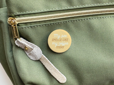 Onderkast Studio’s discontinued “My Pee Smells Like Coffee Today” round yellow pinback button, printed in vibrant colors, is shown on a green fabric bag with a gold zipper and a suede zipper pull.