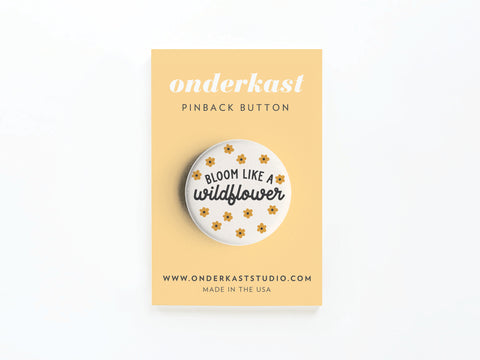 The Bloom Like a Wildflower Pinback Button by Onderkast Studio features small yellow flowers and the phrase “Bloom Like a Wildflower,” symbolizing optimism and strength. It’s attached to a yellow Onderkast Studio card marked MADE IN THE USA.