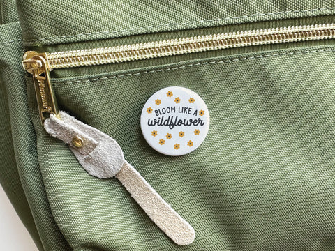 The Bloom Like a Wildflower Pinback Button by Onderkast Studio, featuring yellow flower designs and an optimistic message, is attached to a green fabric bag with a gold zipper and suede pull.