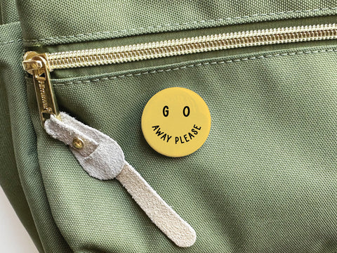 The Onderkast Studio Go Away Please Pinback Button is a round yellow pin that reads GO AWAY PLEASE with two small eye-like dots above the text, perfect for personal space protection.