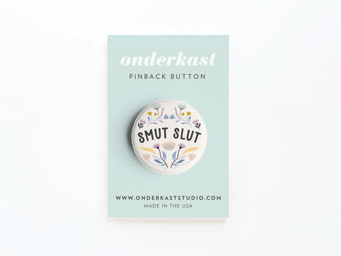 The Smut Slut Pinback Button by Onderkast Studio features literary humor with SMUT SLUT surrounded by floral illustrations, presented on a branded card. Made in the USA by onderkaststudio.com.