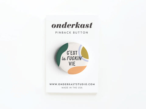 The C’est La Fuckin’ Vie Pinback Button by Onderkast Studio features bold text and abstract shapes, attached to a branded white card showing the Onderkast Studio name and website, set against a plain background.