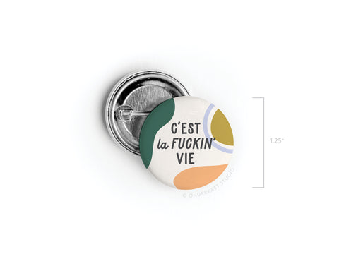 The C’est La Fuckin’ Vie Pinback Button by Onderkast Studio is a round 1.25-inch button with French flair, bold abstract green, yellow, and blue shapes, and C’EST la FUCKIN’ VIE in black on white. Shown facing up and slightly open.