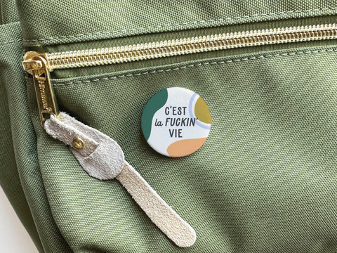 The C’est La Fuckin’ Vie Pinback Button by Onderkast Studio brings French flair to any green fabric bag, featuring bold black lettering on pastel shapes and a stylish gold zipper accent.