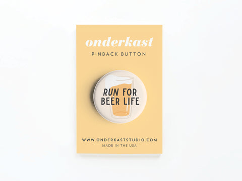 The Run For Beer Life Pinback Button by Onderkast Studio features a beer glass and bold text, displayed on a yellow onderkast card with their website. Made in the USA—perfect for beer enthusiasts!.
