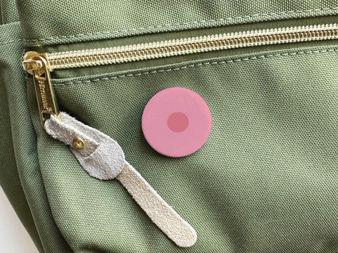 The Nipple Pinback Button by Onderkast Studio—a pink circular pin with a darker center—is shown attached to a light green fabric bag near a gold zipper and beige suede pull tab. This item is discontinued.