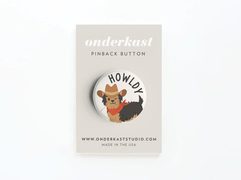 The Howldy Pinback Button by Onderkast Studio features a dapper dog in a cowboy hat and red bandana with HOWLDY above, displayed on an Onderkast Studio card. This Western-inspired accessory adds charm to any outfit.