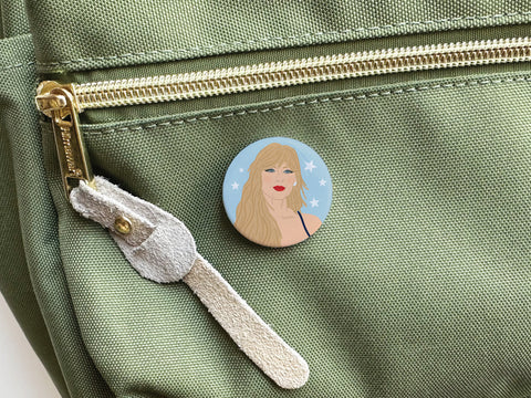 The TS Midnights Pinback Button by Onderkast Studio, featuring an illustrated blonde woman on a blue celestial background, is pinned to the front pocket of a light green fabric bag with a gold zipper and beige suede pull tab.