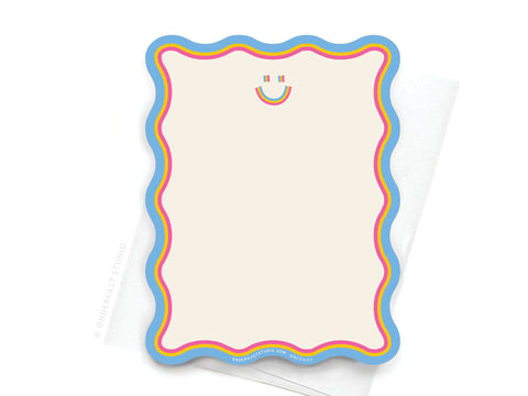 The Miss Ya Wavy Die Cut Notecard by Onderkast Studio features blue, pink, and yellow borders with a multicolored smiley face, resting on a white folded napkin. Crafted from premium card stock for durability.
