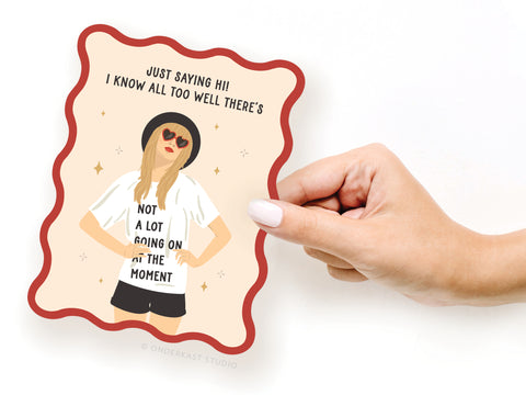 A hand holds the “TS Red Wavy Die Cut Notecard” by Onderkast Studio, featuring a pop culture illustration of a woman in sunglasses, hat, and shorts. Her shirt says “NOT A LOT GOING ON AT THE MOMENT.” The card reads: “JUST SAYING HI!”.