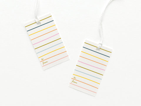 The Bright Lines Gift Tag by Onderkast Studio features two tags with colorful horizontal stripes, to: and from: in gold at the bottom left, and each tag attached to a white string, all shown on a white background.