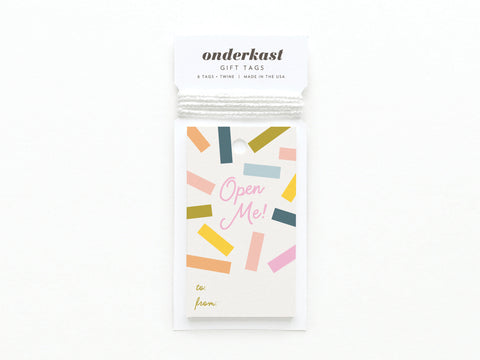 The Open Me Confetti Gift Tag set by Onderkast Studio features six vibrantly printed tags with rectangular confetti, “Open Me!” in pink script, “to:/from:” fields, twine, and sturdy card stock. Made in the USA.