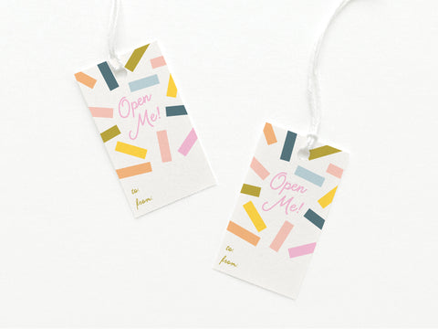 The Open Me Confetti Gift Tag by Onderkast Studio includes two sturdy white tags with vibrant confetti, “Open Me!” in pink, gold to/from fields, and a sleek white string attached.