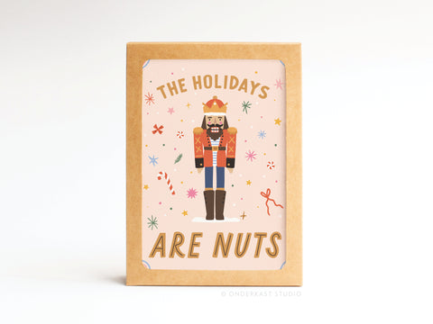The Holidays Are Nuts Nutcracker Folded Greeting Note Set of 10 by Onderkast Studio features bold cartoon nutcrackers and festive decor on a pink background, with vibrant premium printing and includes a brown holder.