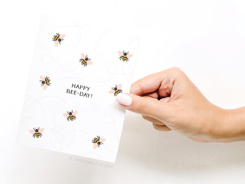 A hand with light pink nail polish holds the Onderkast Studio Happy Bee-day! Greeting Card, featuring cute bees and Happy Bee-day! text on a white background.