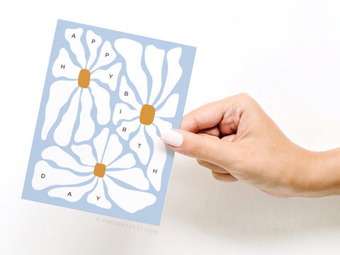 A hand with white nail polish holds the Onderkast Studio Happy Birthday Groovy Flowers Greeting Card—a blue card featuring playful, vibrant flowers, with each petal spelling out Happy Birthday.