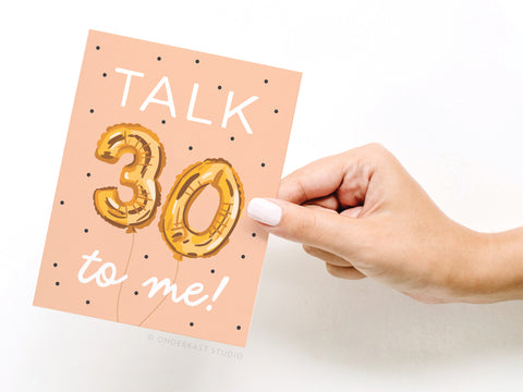 A hand with white nail polish holds the Onderkast Studio Talk 30 to Me Balloons Greeting Card, a peach card with black dots and gold foil balloon-style 30—perfect for celebrating a milestone birthday.
