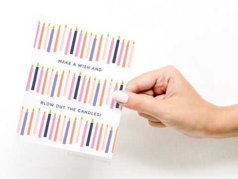 A hand with light pink nail polish holds the Onderkast Studio Make a Wish and Blow Out the Candles Greeting Card, featuring colorful rows of birthday candles and the phrase “MAKE A WISH AND BLOW OUT THE CANDLES!” on a white background.