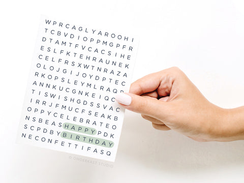 A hand with pale pink nails holds the Onderkast Studio Happy Birthday Word Search Greeting Card, featuring “HAPPY BIRTHDAY” highlighted in green within a playful black-letter puzzle design.