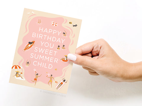 A hand holds the Onderkast Studio Happy Birthday You Sweet Summer Child Greeting Card featuring summer-themed illustrations like people swimming, sunbathing, and playing around the message on a pink background.