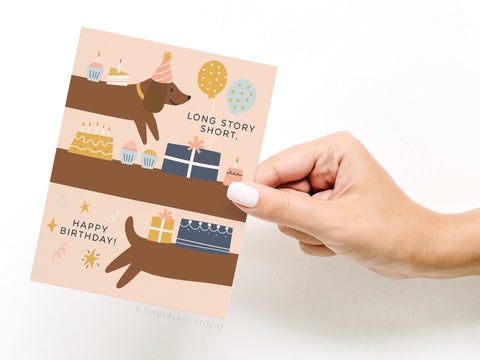 A hand holds the Onderkast Studio Long Story Short Dog Birthday Greeting Card featuring a cartoon dachshund in a party hat with cakes, cupcakes, balloons, and presents. The card reads “LONG STORY SHORT, HAPPY BIRTHDAY!”.